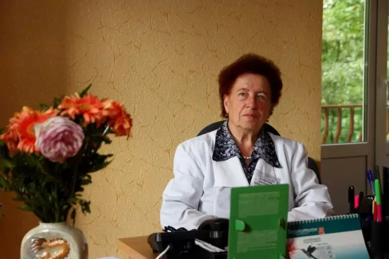 Женщина в белом медицинском халате сидит за рабочим столом в кабинете, на переднем плане цветы, а на заднем фоне окно с видом на зелень